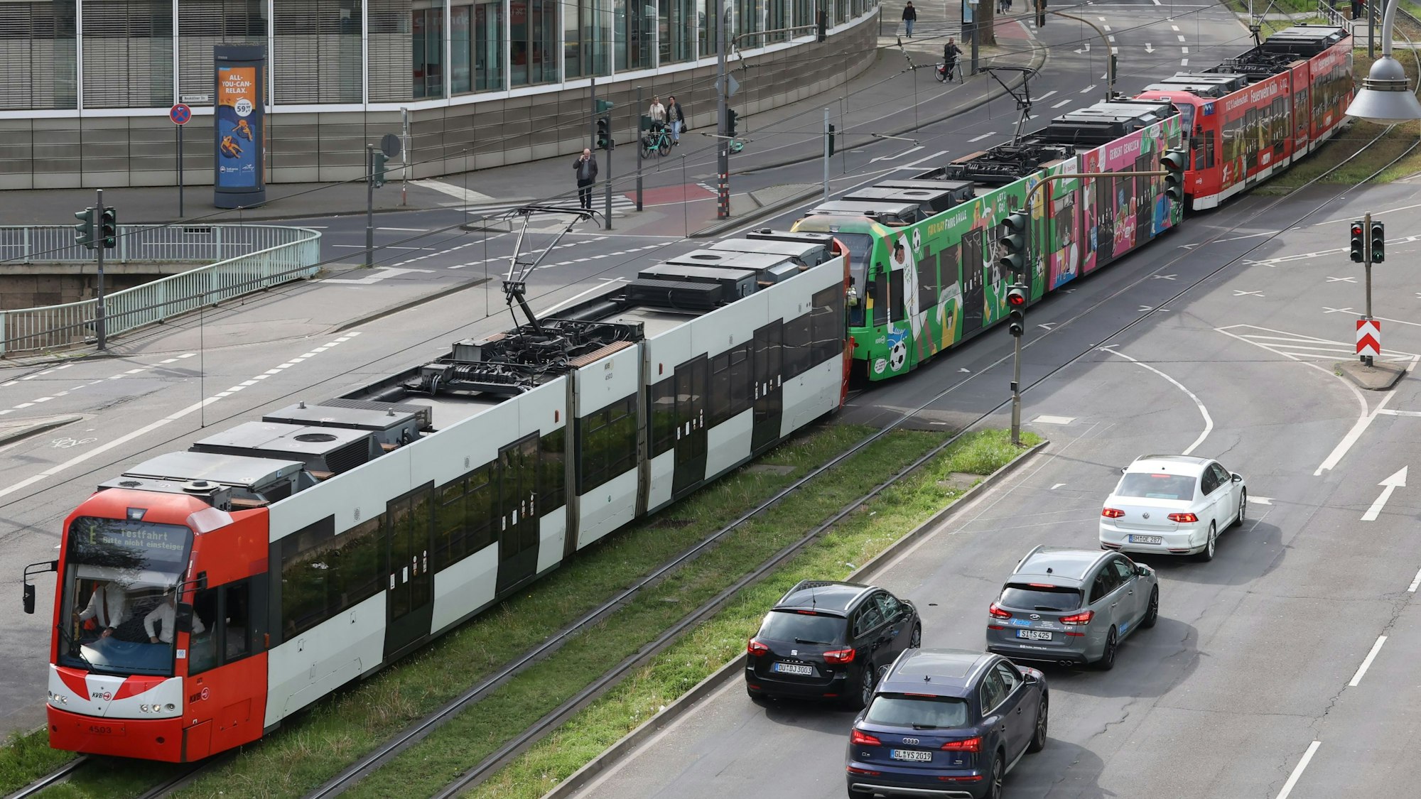 Ein aus drei Straßenbahnen bestehender Langzug befährt das Gleisbett auf der Kölner Cäcilienstraße. Autos, die links abbiegen wollen, warten vor einer Ampel auf die Durchfahrt der 90-Meter-Bahn.