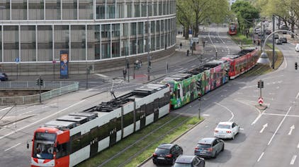 Ein aus drei Straßenbahnen bestehender Langzug befährt das Gleisbett auf der Kölner Cäcilienstraße. Autos, die links abbiegen wollen, warten vor einer Ampel auf die Durchfahrt der 90-Meter-Bahn.&nbsp;