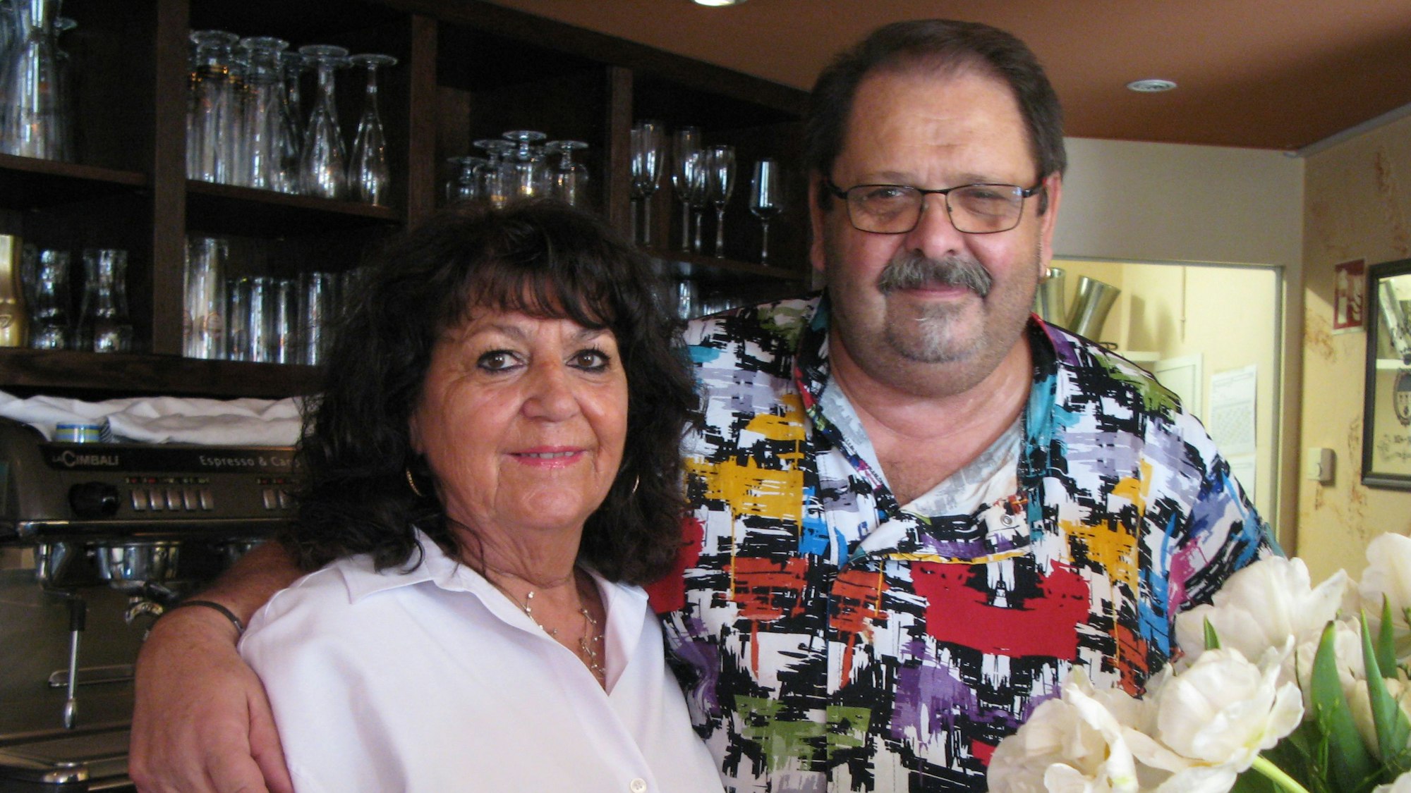 Das Ehepaar Manuela und Bruno Weber steht hinter dem Tresen. Sie trägt eine weiße Bluse, er ein buntgemustertes Hemd.