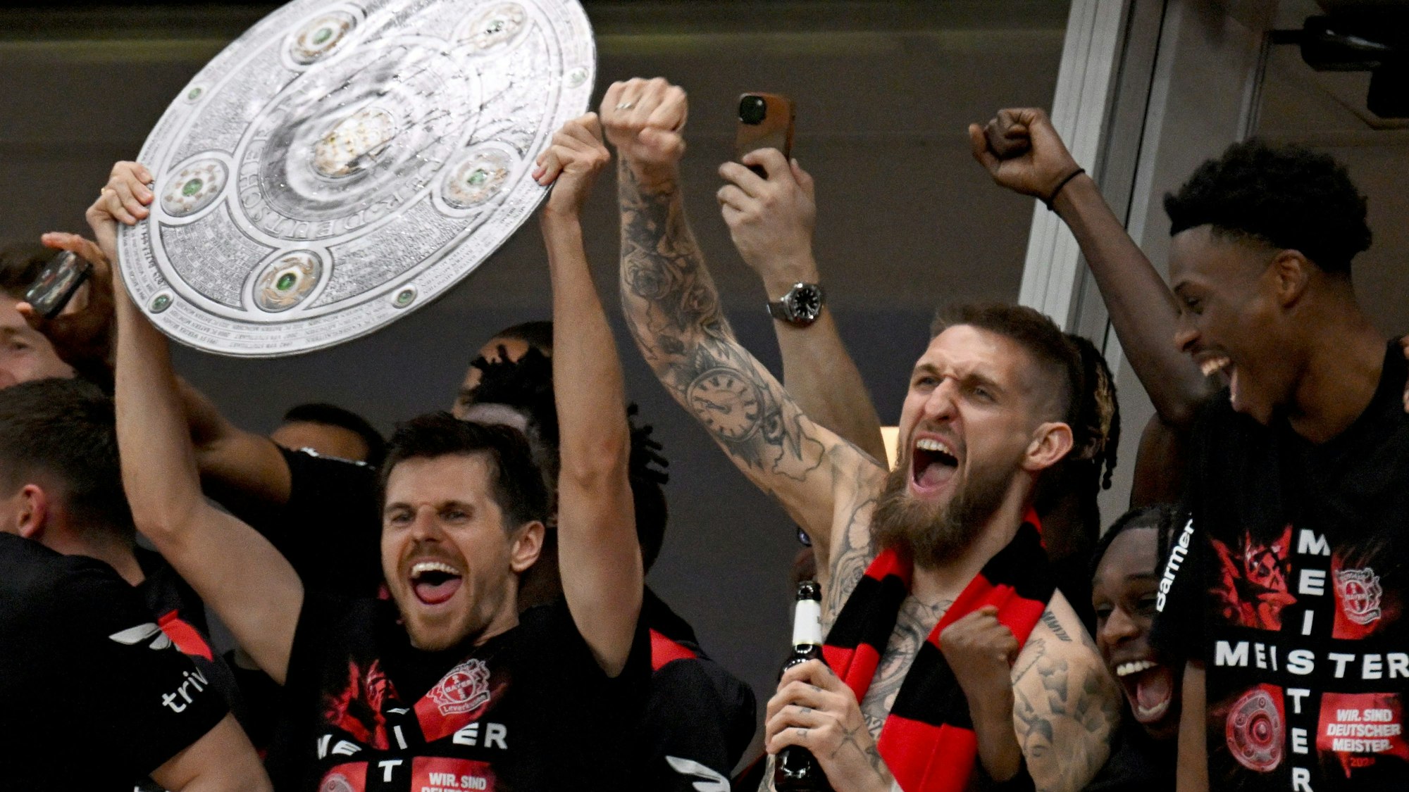 Die Leverkusener Mannschaft jubelt mit einer stilisierten Meisterschale von der Tribüne ins Stadion, den Fans zu, die nach dem Sieg das Spielfeld gestürmt haben.