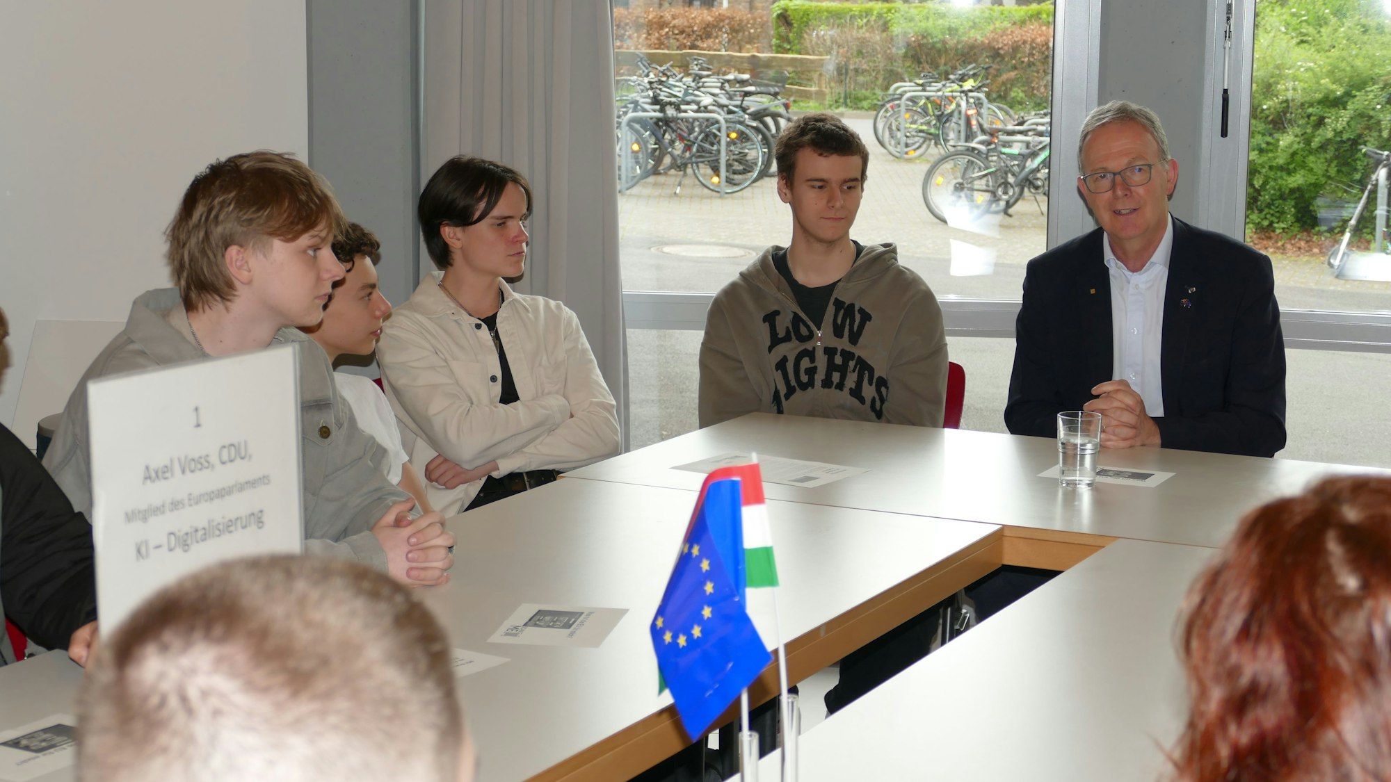 Jugendliche und ein Mann mit grauen Haaren sitzen an einem Tisch, auf dem ein EU-Fähnchen steht.