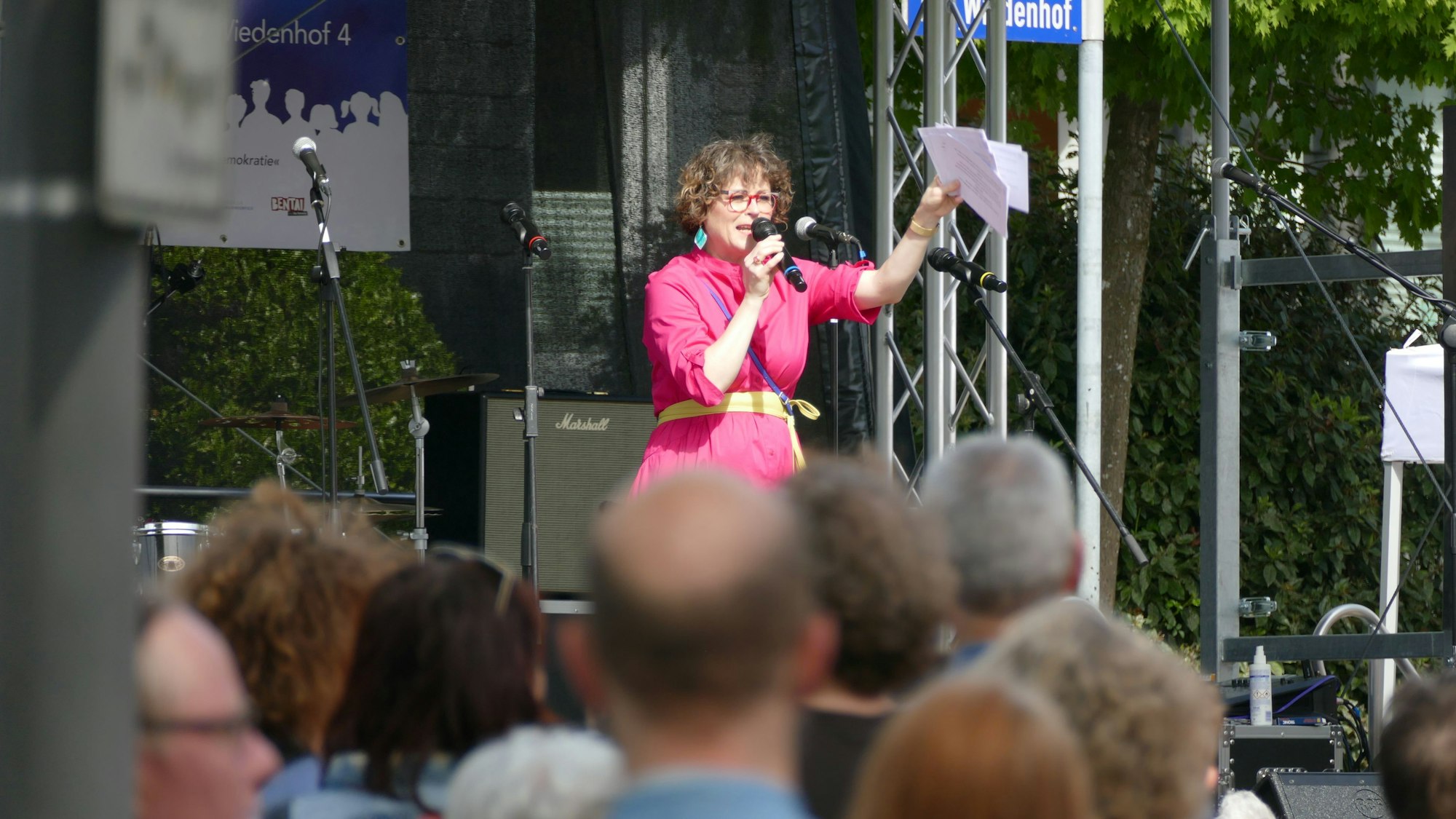 Eine Frau mit pinkfarbenem Kleid steht auf einer Bühne und spricht in ein Mikrofon.