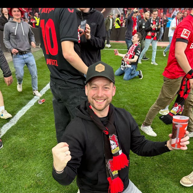 Ein Mann mit Fan-Schal von Bayer 04 Leverkusen strahlt und reckt die Faust in die Höhe. Er steht auf dem Spielfeld.&nbsp;
