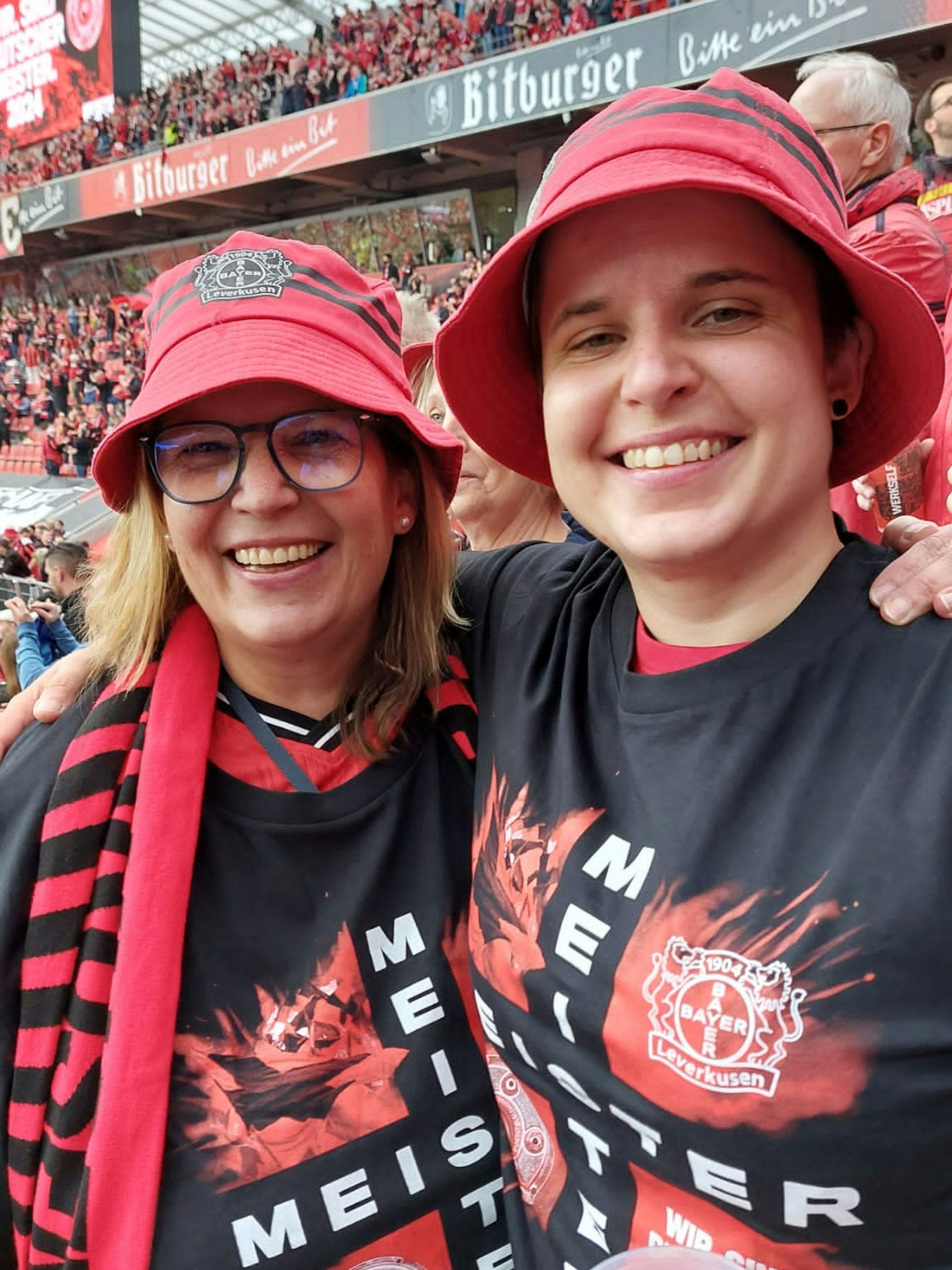 Zwei Frauen mkt roten Fan-Hüten und schwarzen T-Shirts von Bayer 04, auf denen „Meister“ steht.