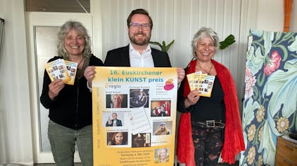 Gaby Bärenklau und Heike Jachtmann halten das Programmheft des 16. Euskirchener Kleinkunstpreises in den Händen. Sie flankieren Sebastian Zimer, der das zugehörige Plakat präsentiert.