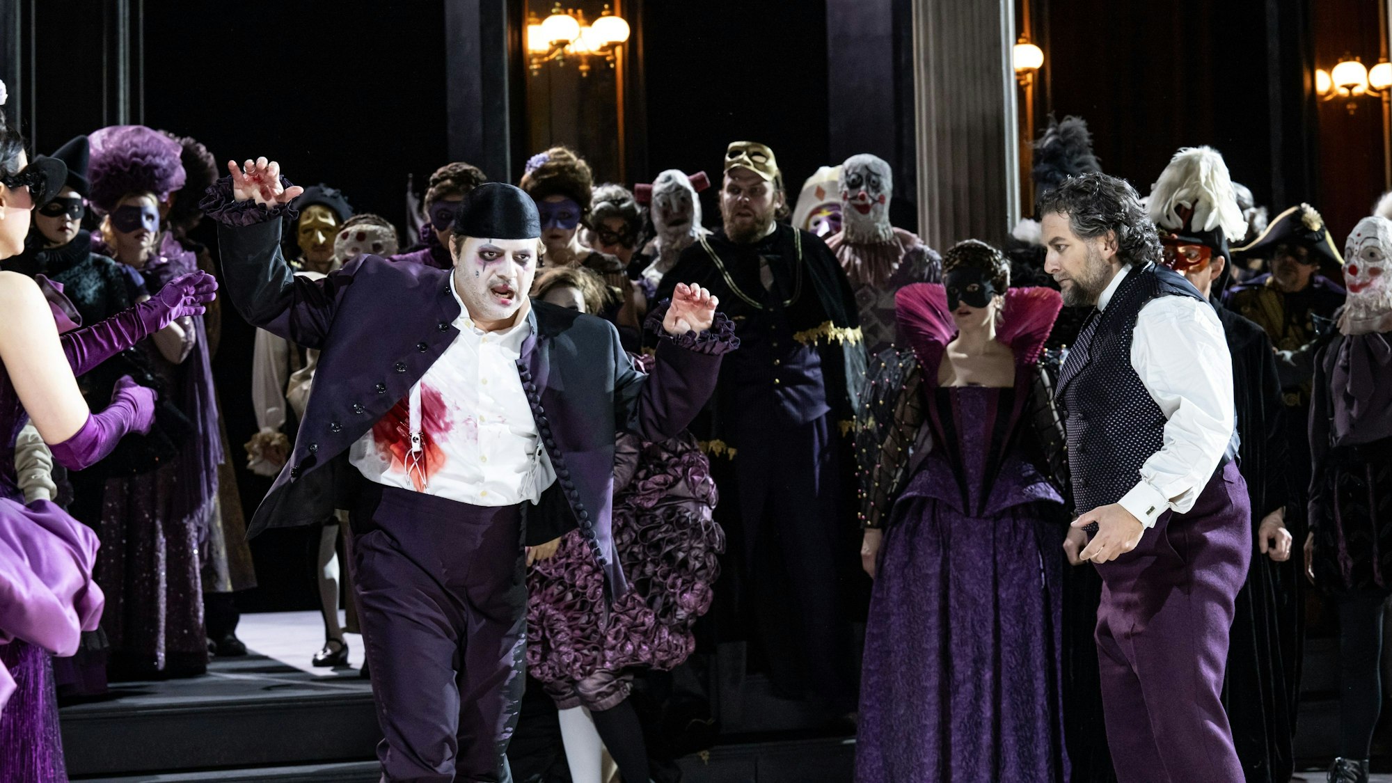 Riccardo wirkt mit weiß geschminktem Gesicht und violettem Anzug ein bisschen wie der Joker. Neben ihm Simone del Savio, ebenfalls in violetter Hose, aber weißem Hemd und einer Weste. Im Hintergrund das bunte Treiben des Chores der Kölner Oper.