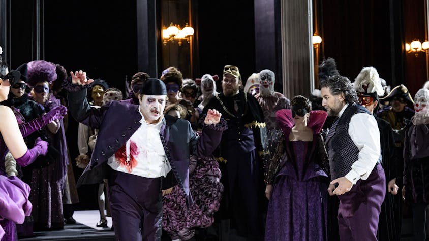 Riccardo wirkt mit weiß geschminktem Gesicht und violettem Anzug ein bisschen wie der Joker. Neben ihm Simone del Savio, ebenfalls in violetter Hose, aber weißem Hemd und einer Weste. Im Hintergrund das bunte Treiben des Chores der Kölner Oper.&nbsp;