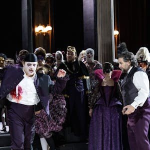 Riccardo wirkt mit weiß geschminktem Gesicht und violettem Anzug ein bisschen wie der Joker. Neben ihm Simone del Savio, ebenfalls in violetter Hose, aber weißem Hemd und einer Weste. Im Hintergrund das bunte Treiben des Chores der Kölner Oper.&nbsp;