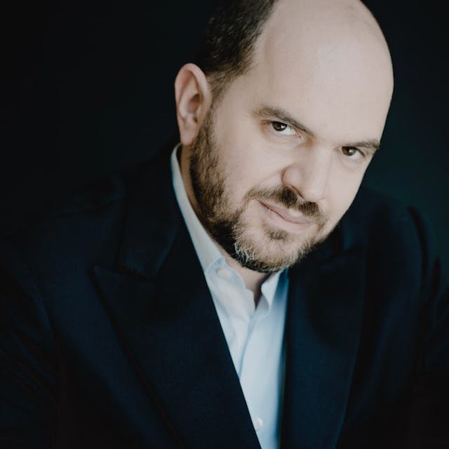 Auf dem Bild ist der Pianist Kirill Gerstein zu sehen. Er trägt einen grau melierten Bart und eine Halbglatze. Er trägt einen dunklen Anzug und ein hellblaues Hemd.