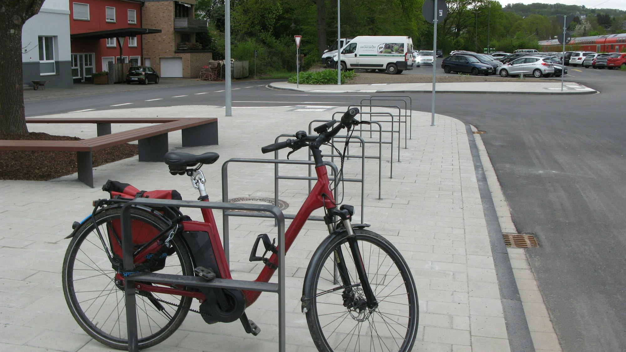 Ein rotes Fahrrad ist einem Anlehnbügel abgestellt, um einen Baum wurde eine Sitzbank installiert. Im Hintergrund ist der Park-and-Ride-Platz zu sehen, auf dem ein Parkhaus gebaut werden soll.