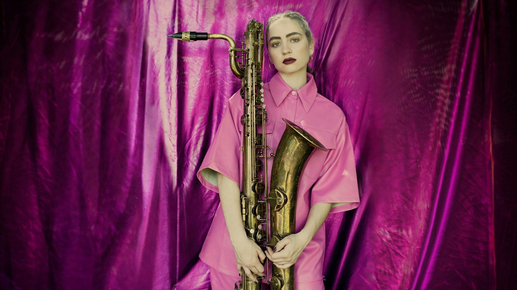 Auf dem Bild ist eine blonde Frau in einem pinken Hemd zu sehen. Sie hält ein Saxophon. Im Hintergrund ist ein glänzender, pinker Vorhang.