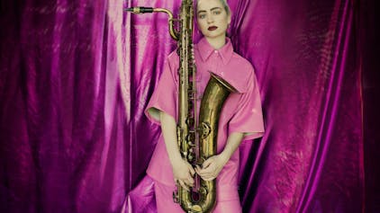 Auf dem Bild ist eine blonde Frau in einem pinken Hemd zu sehen. Sie hält ein Saxophon. Im Hintergrund ist ein glänzender, pinker Vorhang.&nbsp;