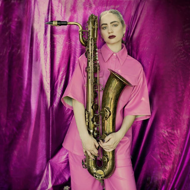 Auf dem Bild ist eine blonde Frau in einem pinken Hemd zu sehen. Sie hält ein Saxophon. Im Hintergrund ist ein glänzender, pinker Vorhang.&nbsp;