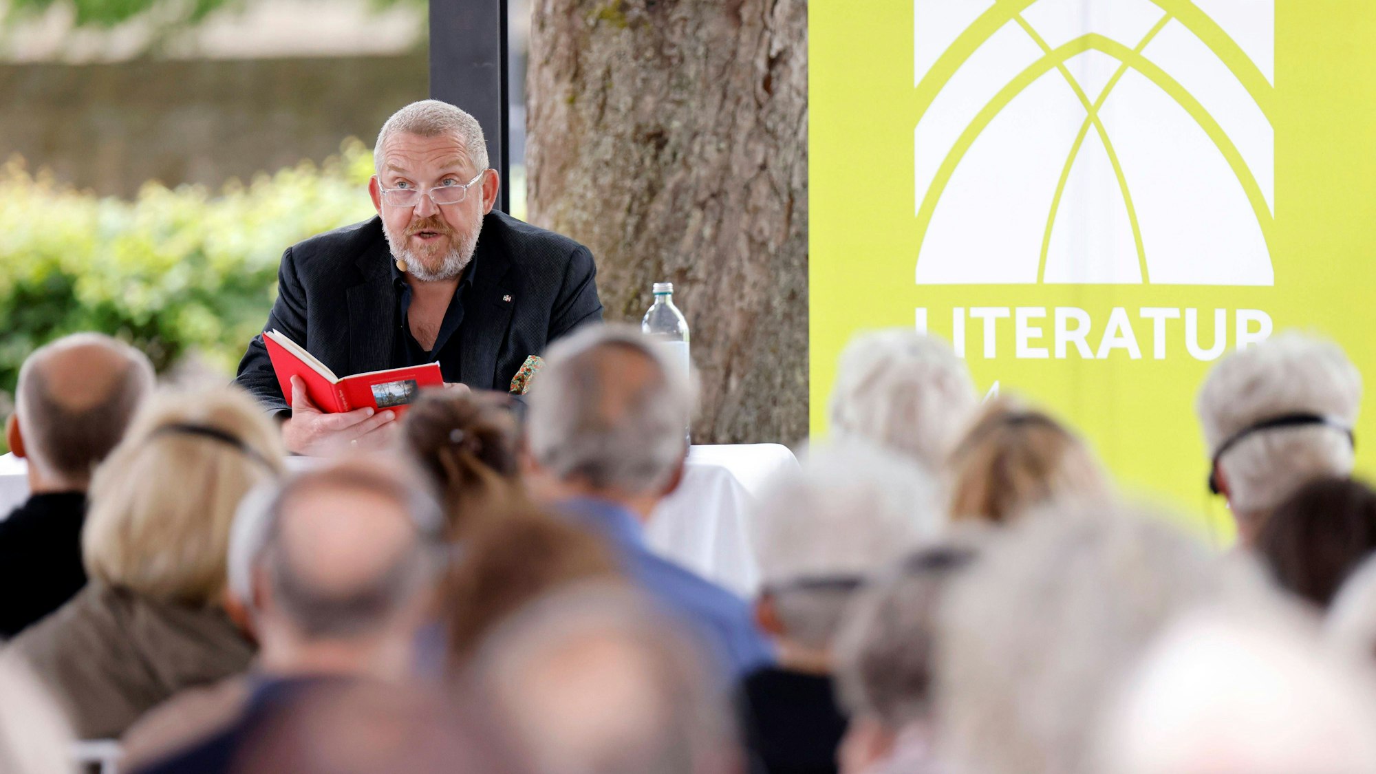 Schauspieler Dietmar Bär mit Lesebrille bei einer Lesung im Rahmen des Literaturfestivals Literatur am Dom am Altenberger Dom. Vor ihm ein verschwommenes Publikum, hinter ihm auf der Bühne ein grünes Banner mit der Aufschrift „Literatur am Dom“.