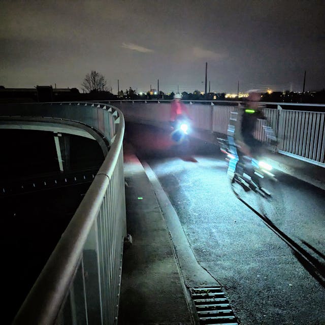 Zwei Radfahrer mit beleuchteten Fahrrädern fahren in der Dunkelheit über eine Brücke.