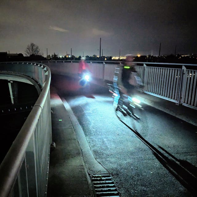 Zwei Radfahrer mit beleuchteten Fahrrädern fahren in der Dunkelheit über eine Brücke.