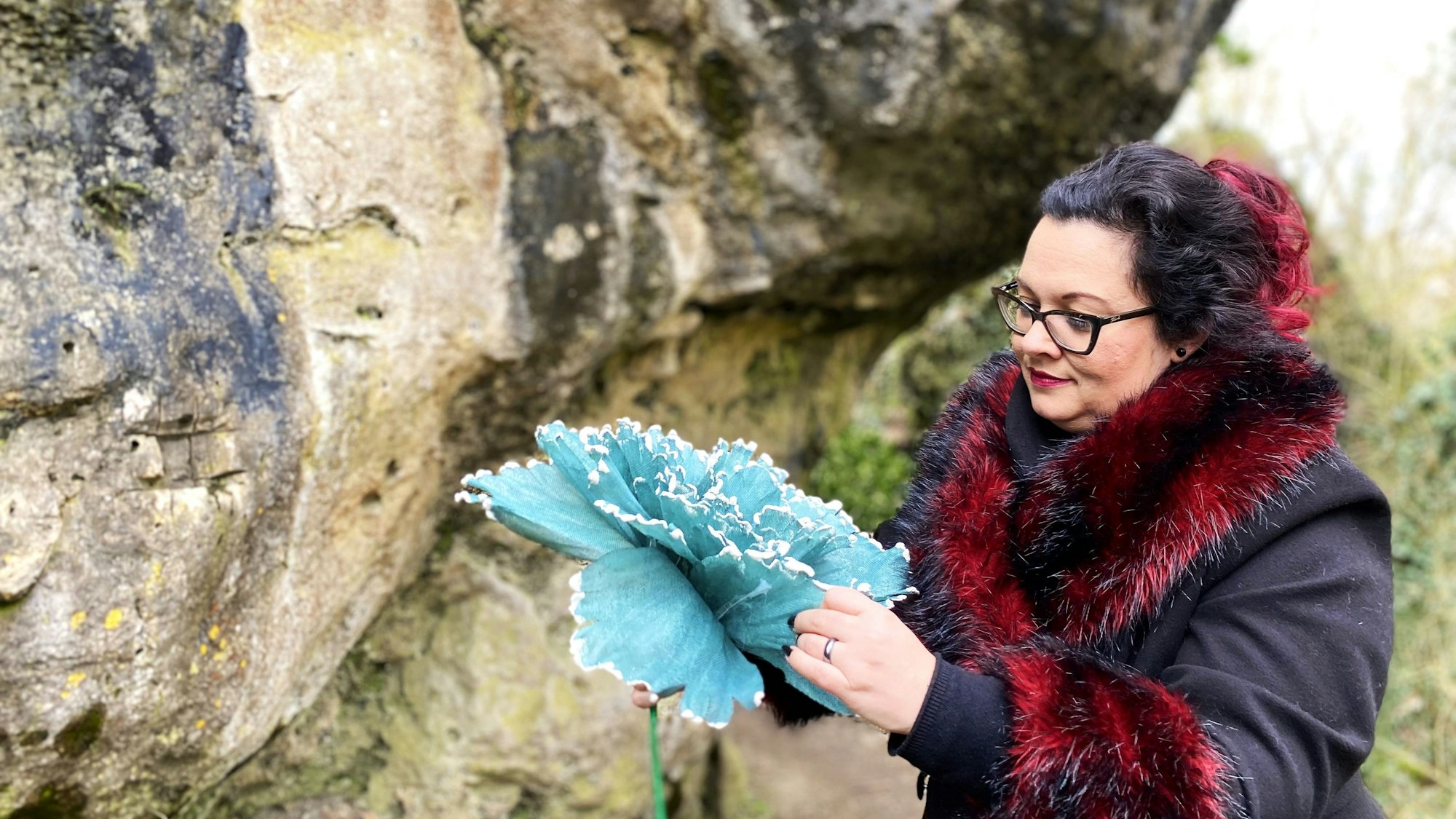 Vanessa Gebhardt steht vor der Kakushöhle in Mechernich und faltet eine selbstgebastelte blaue Blume auf.