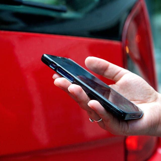 Das Foto zeigt eine Hand mit einem Smartphone und im Hintergrund ein rotes Auto.