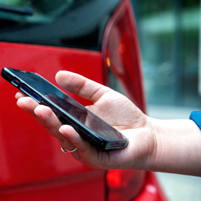 Das Foto zeigt eine Hand mit einem Smartphone und im Hintergrund ein rotes Auto.