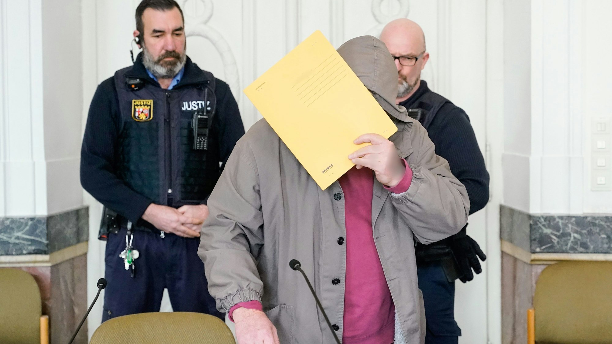 Der Angeklagte steht im Gerichtssaal und verdeckt sein Gesicht mit einer Mappe.