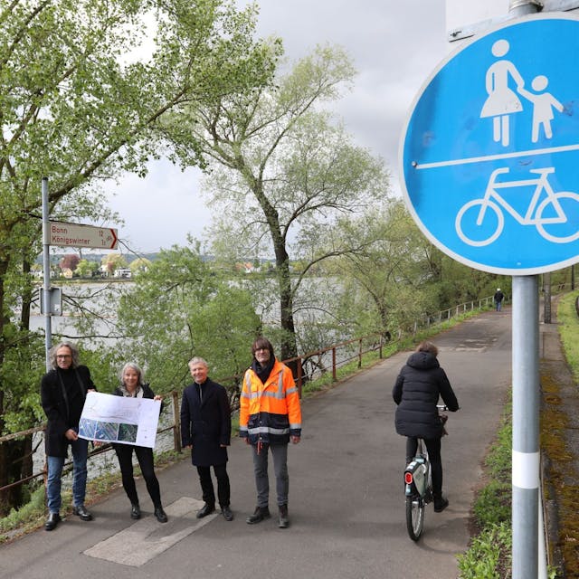 Eine vierköpfige Gruppe steht auf einem Radweg, am Rand weisen rot-weiße Schilder nach Bonn und Königswinter..