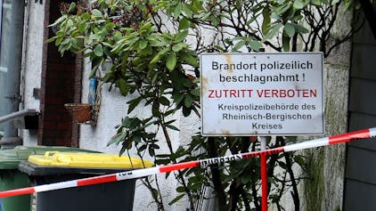 Vor einem Haus steht ein Schild mit der Aufschrift „Brandort polizeilich beschlagnahmt! Zutritt verboten“. Davor ist Polizei-Absperrband zu sehen.