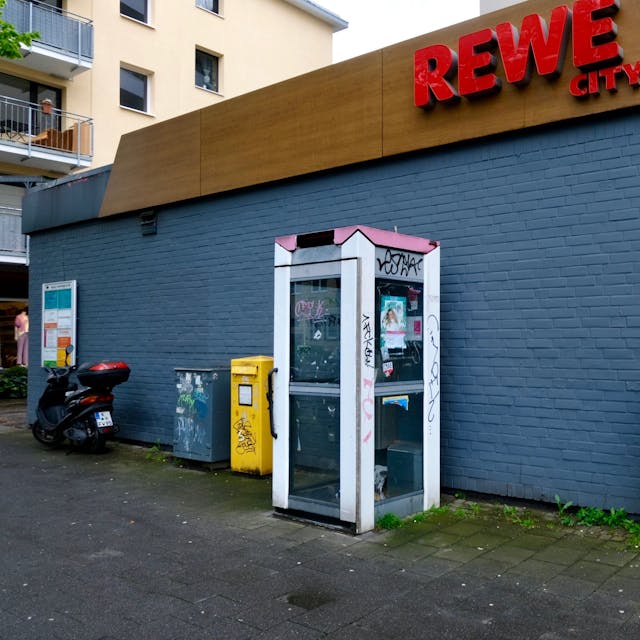 Das Bild zeigt eine verwahrloste und nicht mehr funktionstüchtige Telefonzelle vor dem Rewe-Markt an der Dürener Straße. Foto von Susanne Esch