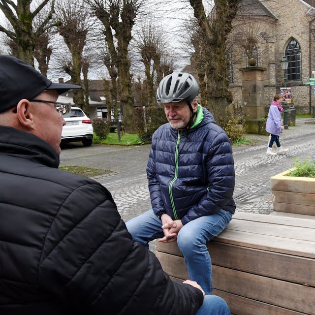 Zwei Männer sitzen auf neuen Bänken an einer Straße in Lindlar.