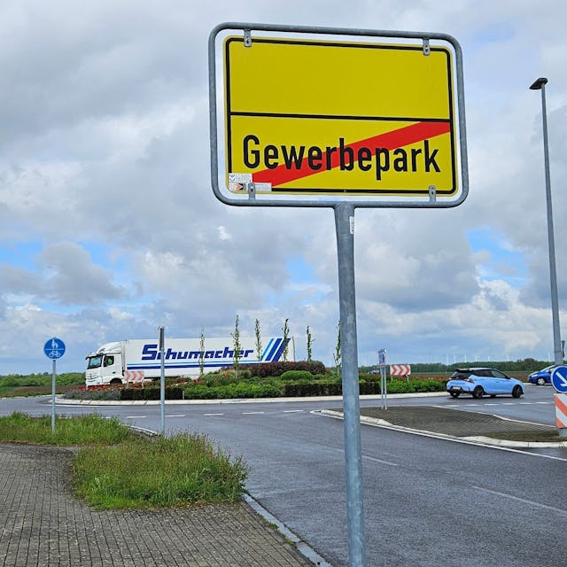 Ein Schild zeigt das Ende eines Gewerbeparks an, dahinter fahren Autos und ein Lastwagen durch einen Verkehrskreisel.