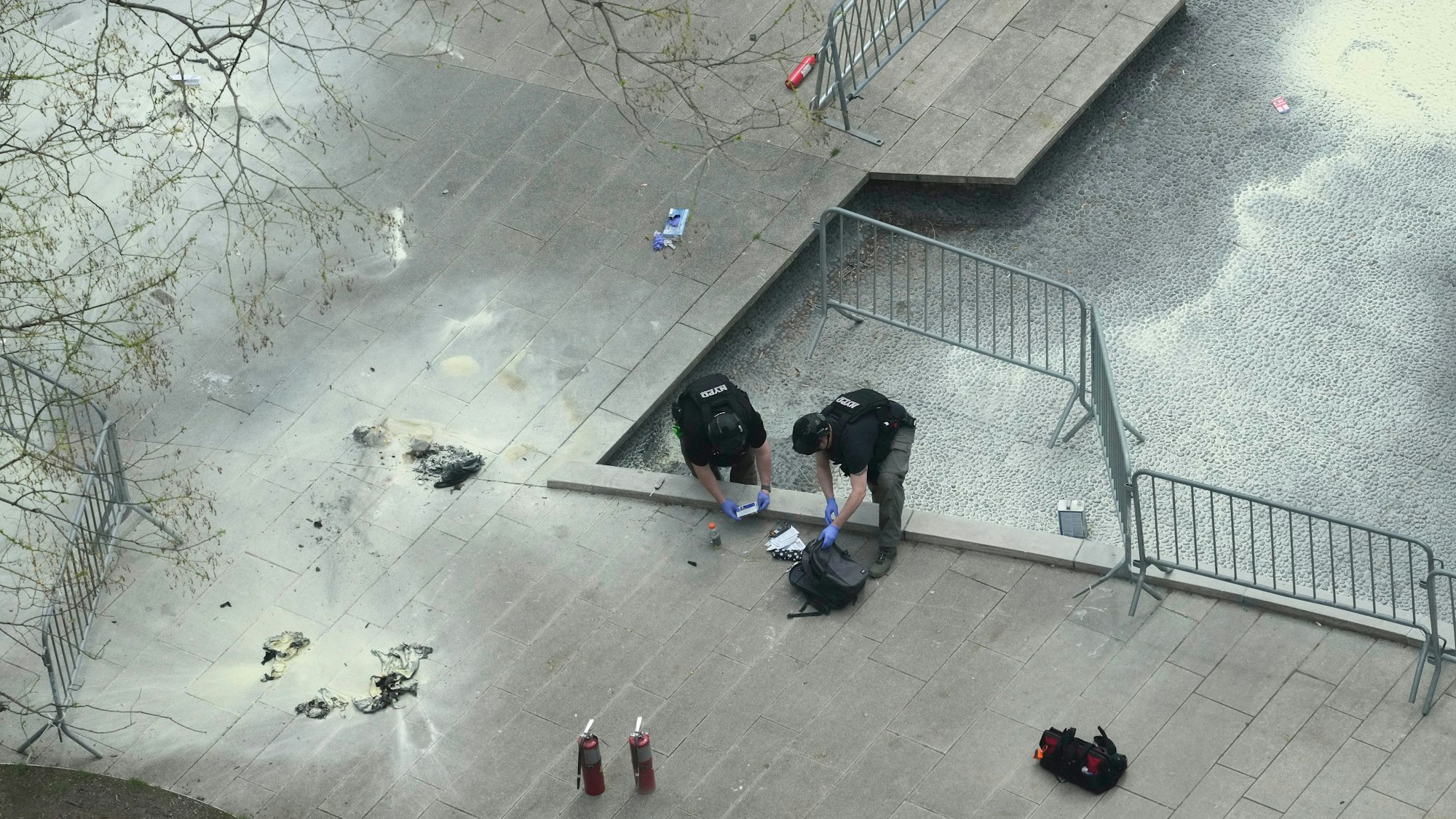 Aufnahme aus der Vogelperspektive auf zwei Polizisten, die mit Handschuhen Gegenstände aus einem Rucksack untersuchen.