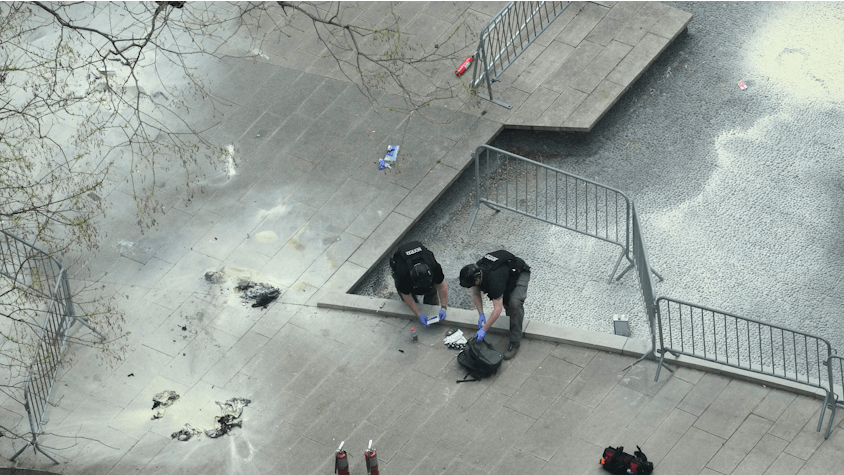 Aufnahme aus der Vogelperspektive auf zwei Polizisten, die mit Handschuhen Gegenstände aus einem Rucksack untersuchen.