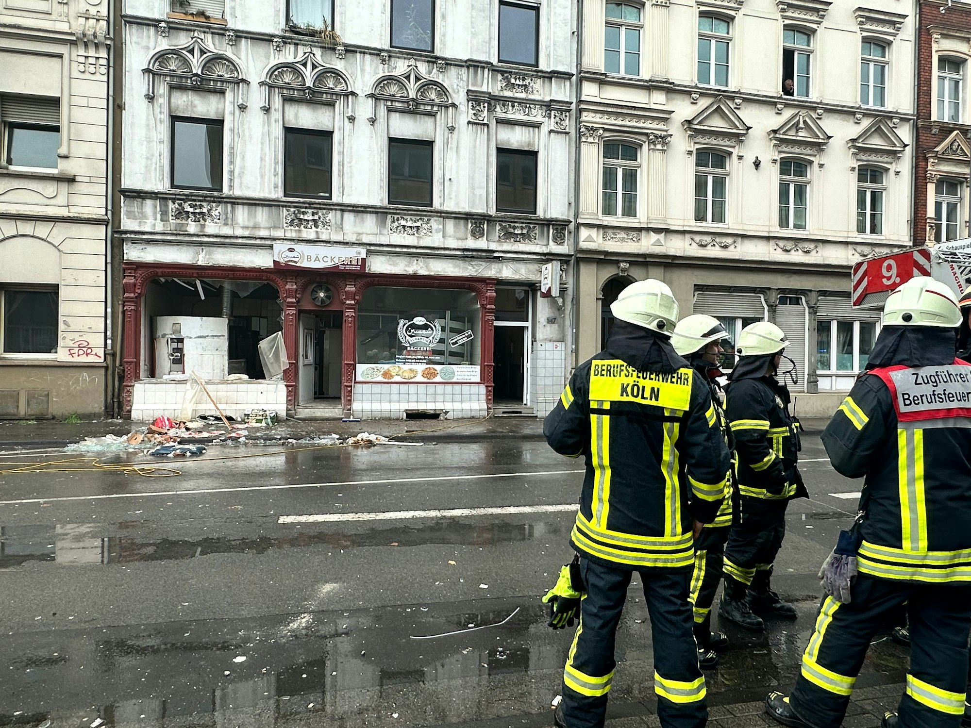 Einsatzkräfte der Feuerwehr Köln stehen vor einer Bäckerei, deren Schaufenster zerborsten sind.