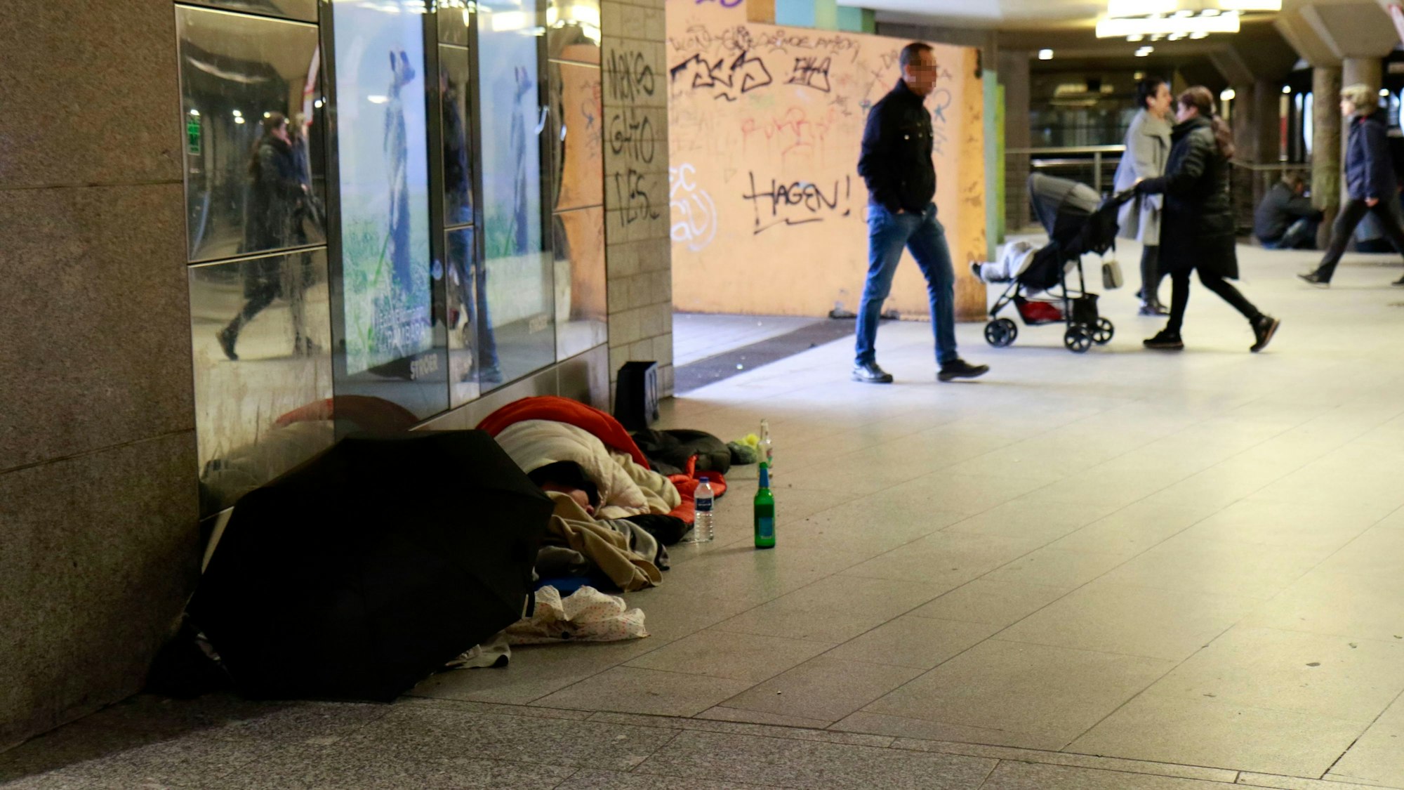 Obdachlose liegen tagsüber am Wiener Platz, im Hintergrund gehen Passanten an ihnen vorbei.