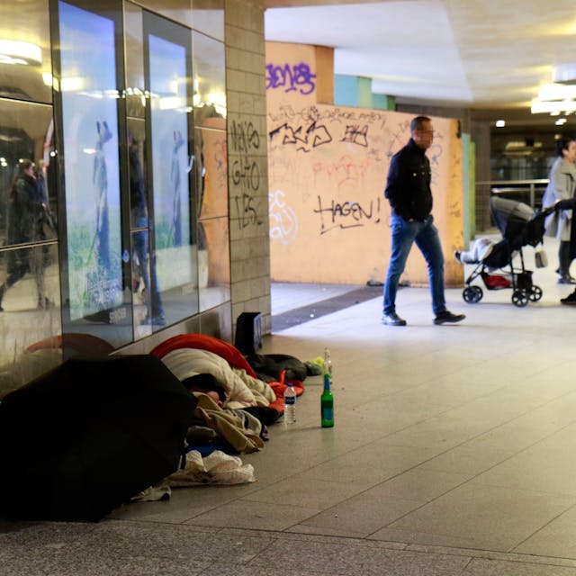 Obdachlose liegen tagsüber am Wiener Platz, im Hintergrund gehen Passanten an ihnen vorbei.