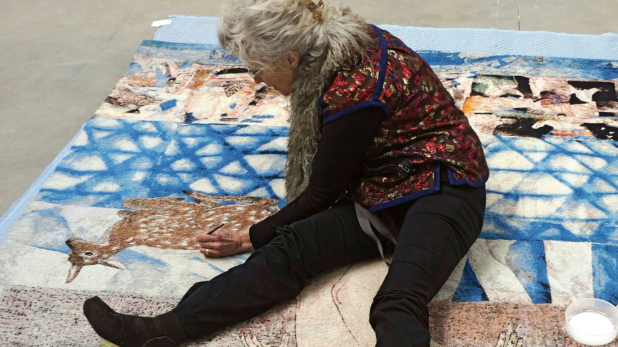 Kiki Smith sitzt auf einer über den Boden ausgebreiteten Papiercollage und malt mit einem Stift.