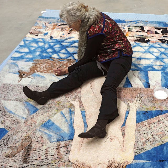 Kiki Smith sitzt auf einer über den Boden ausgebreiteten Papiercollage und malt mit einem Stift.