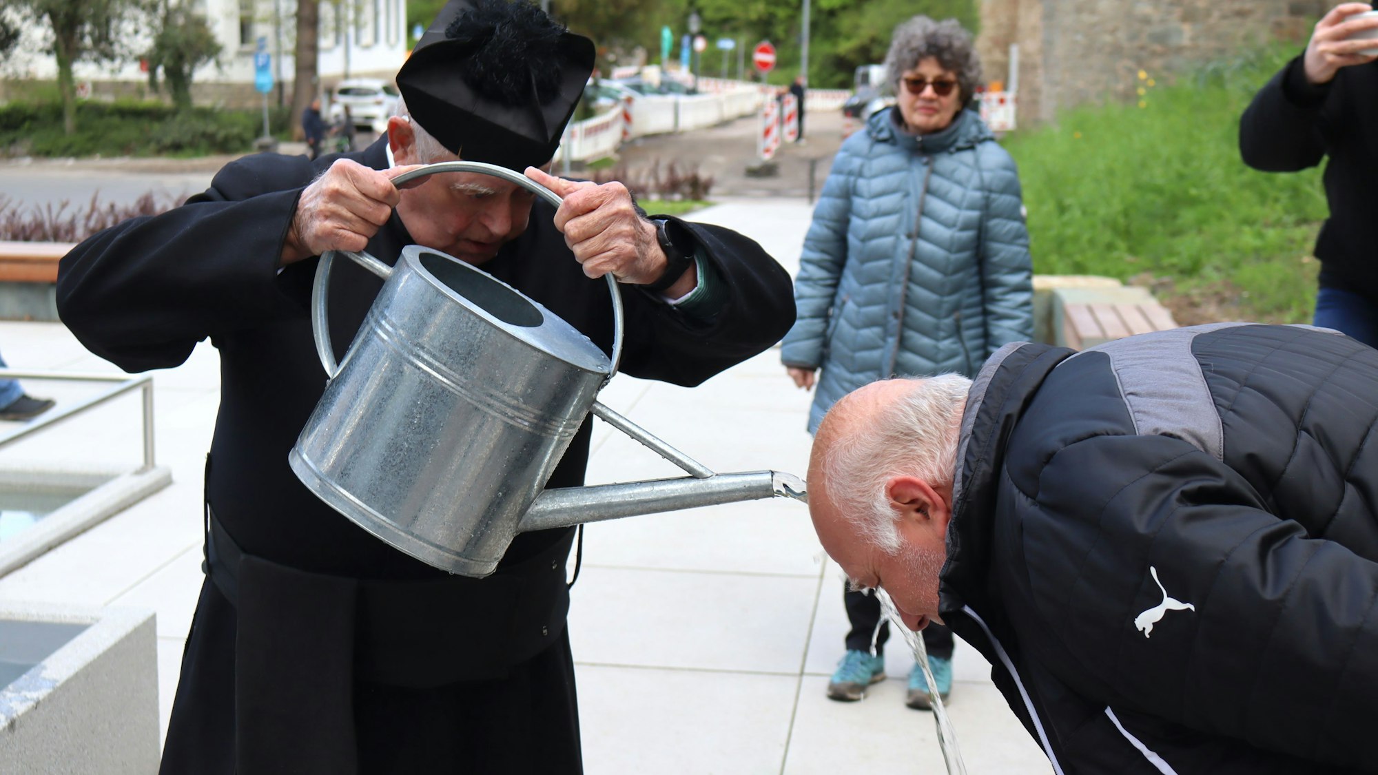 Ein Mann im Pfarrer-Kostüm übergießt einen anderen Mann aus einer Gießkanne mit Wasser.