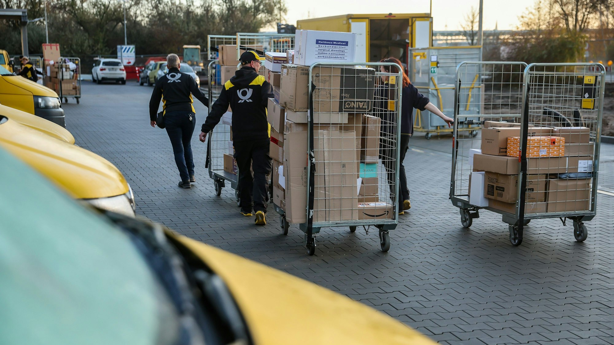 Mitarbeiter und Mitarbeiterinnen der Deutsche Post DHL gehen mit vollen Paketwagen zu ihren Fahrzeug auf dem Hof eines Zustellpunktes. (Archivbild)