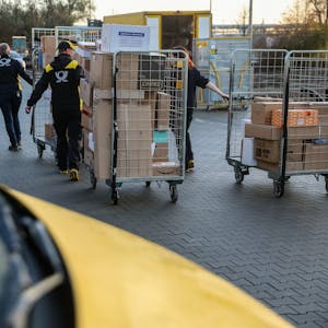 Mitarbeiter und Mitarbeiterinnen der Deutsche Post DHL gehen mit vollen Paketwagen zu ihren Fahrzeug auf dem Hof eines Zustellpunktes. (Archivbild)