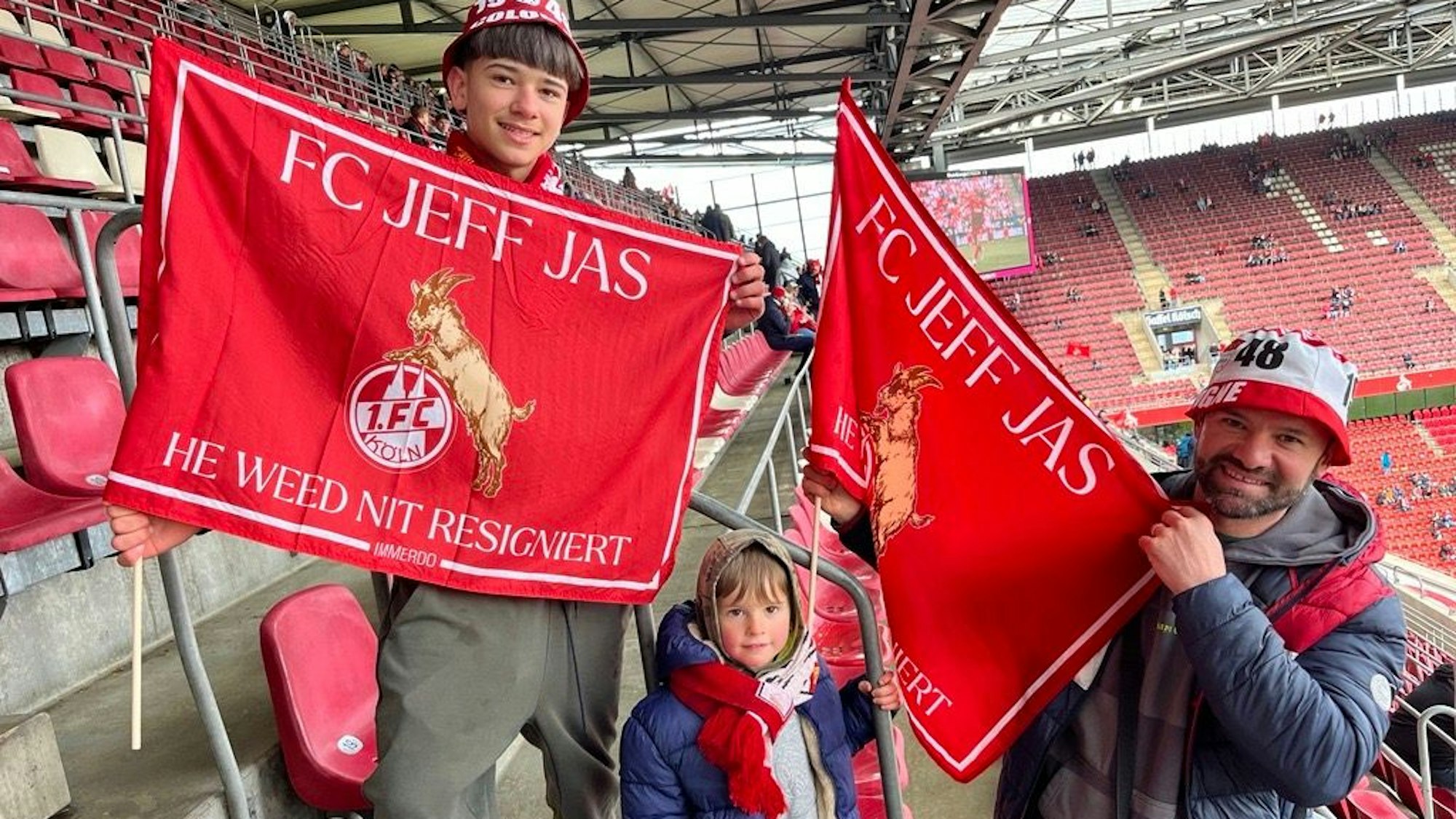Die Kölner Fans Maryo, Noah, und Gigi (v.l.) präsentieren Fahnen mit dem Kölner Rettungs-Motto „FC jeff Jas. He weed nit resigniert“, vor dem Spiel des 1. FC Köln gegen Darmstadt 98.
