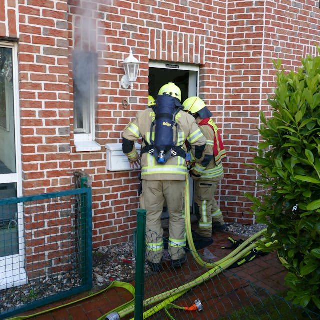 Feuerwehrleute stehen vor einer Haustür, aus dem Fenster daneben kommt Qualm.
