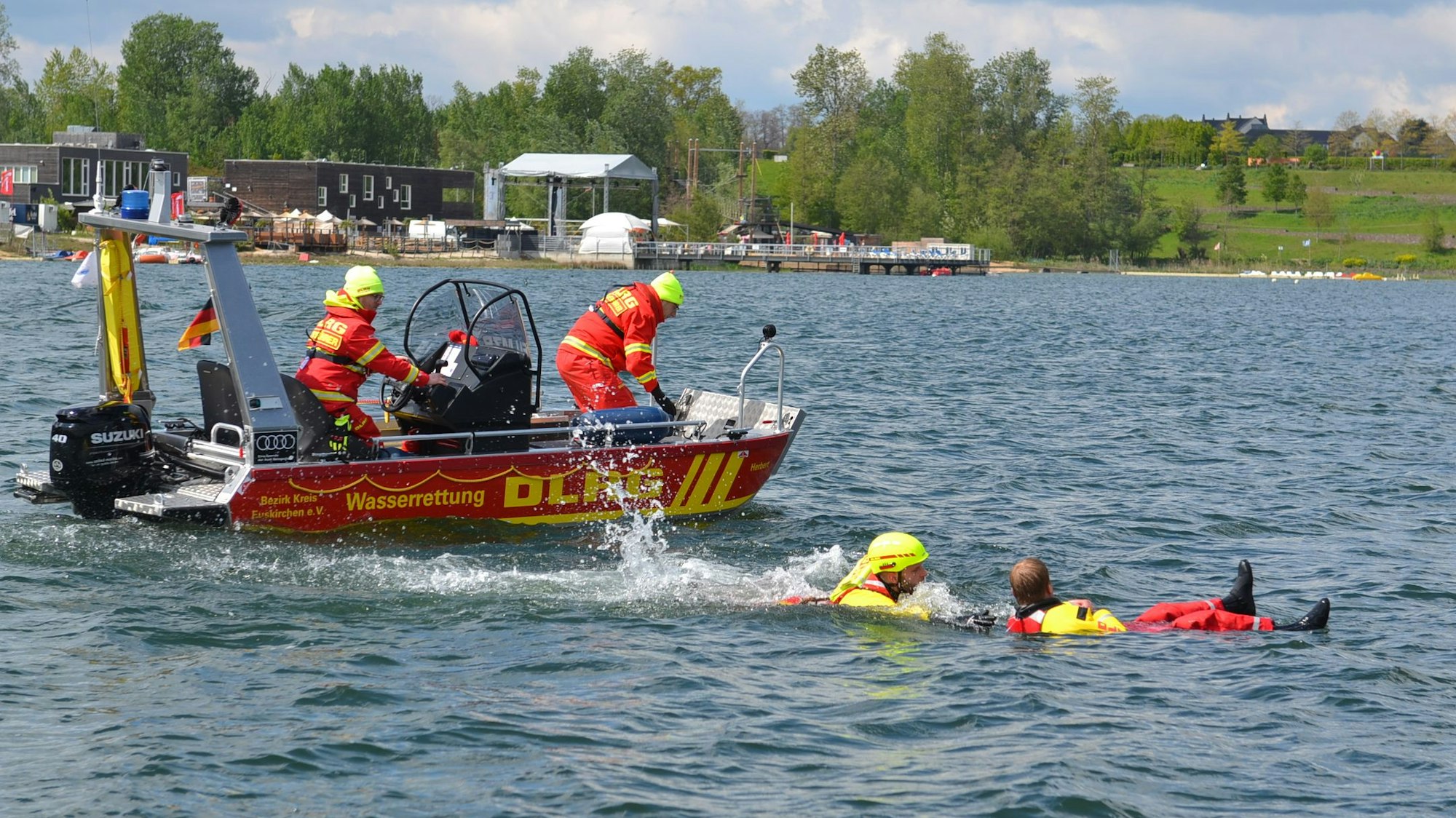 Zwei Einsatzkräfte bedienen das rote Motorboot. Ein dritter DLRG-Helfer ist in den Zülpicher See gesprungen und zieht einen Mann an Bord, der auf dem Rücken im Wasser treibt.
