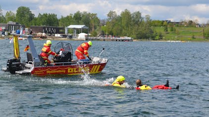 Zwei Einsatzkräfte bedienen das rote Motorboot. Ein dritter DLRG-Helfer ist in den Zülpicher See gesprungen und zieht einen Mann an Bord, der auf dem Rücken im Wasser treibt.