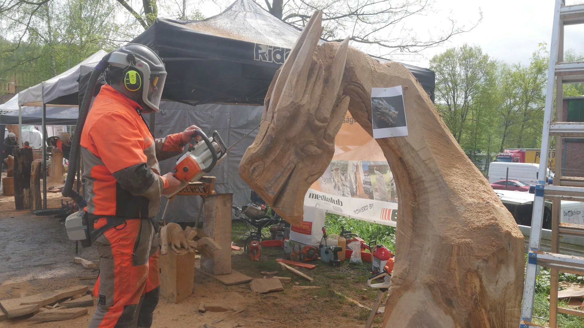 Ein Mann im Schutzanzug und mit Helm arbeitet mit der Kettensäge an einer Holzskulptur.