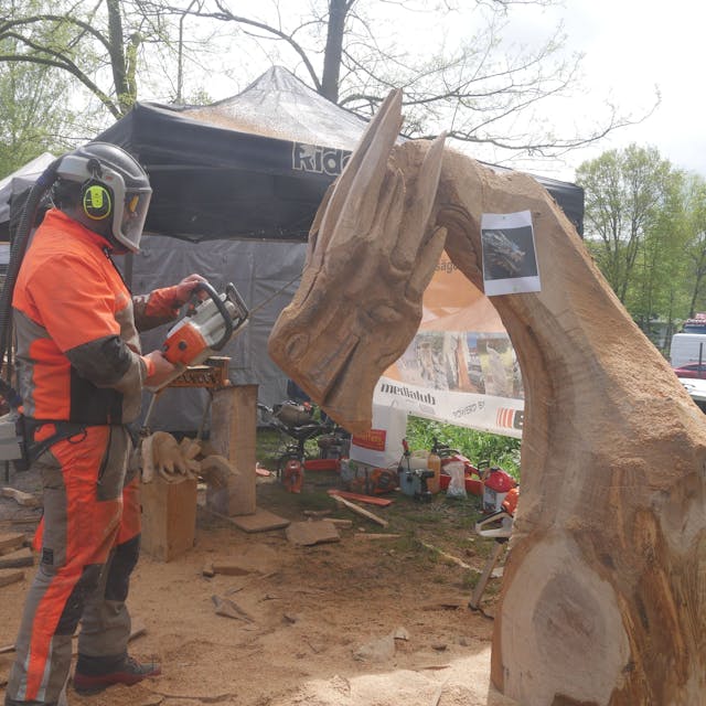 Ein Mann im Schutzanzug und mit Helm arbeitet mit der Kettensäge an einer Holzskulptur.