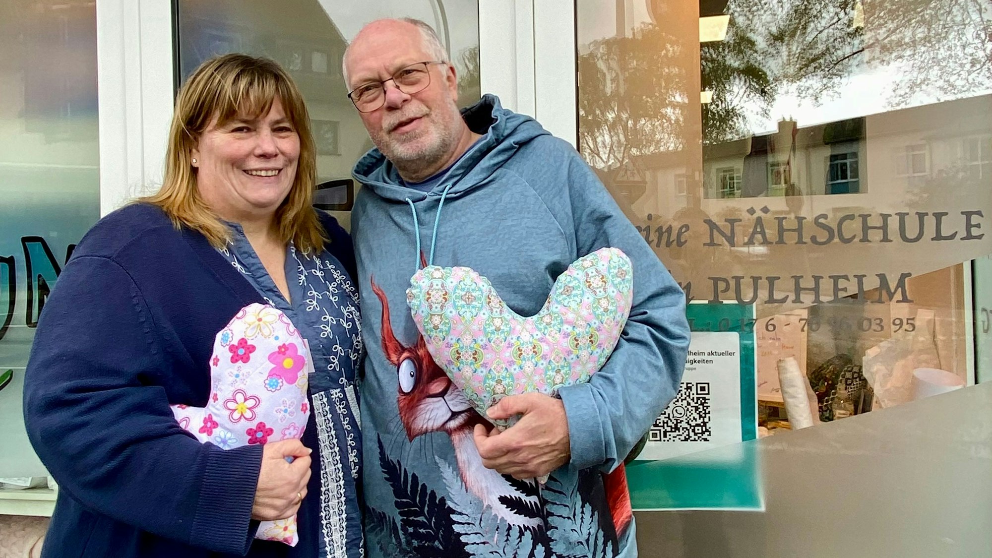 Auf dem Foto sind Heike Baier-Lücke und Thomas Lücke zu sehen. Sie haben den Kölner Herzkissen Verein gegründet, um mit Herzkissen Brustkrebspatientinnen zu unterstützen. Sie halten Herzkissen in der Hand.