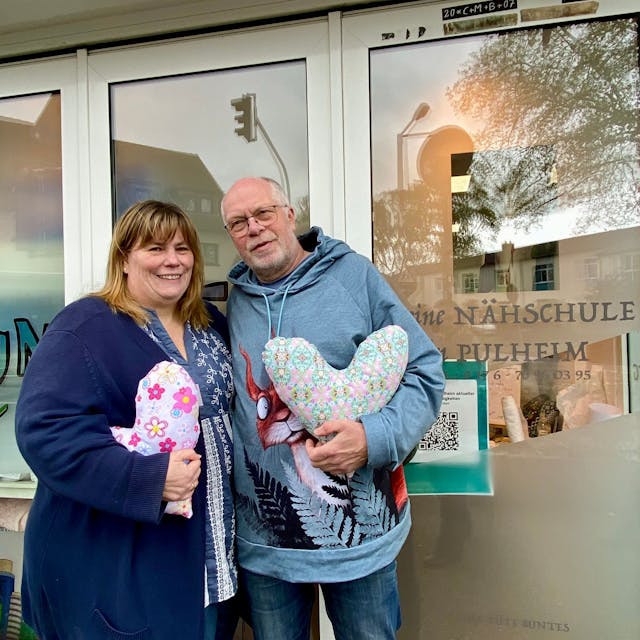 Auf dem Foto sind Heike Baier-Lücke und Thomas Lücke zu sehen. Sie haben den Kölner Herzkissen Verein gegründet, um mit Herzkissen Brustkrebspatientinnen zu unterstützen. Sie halten Herzkissen in der Hand.