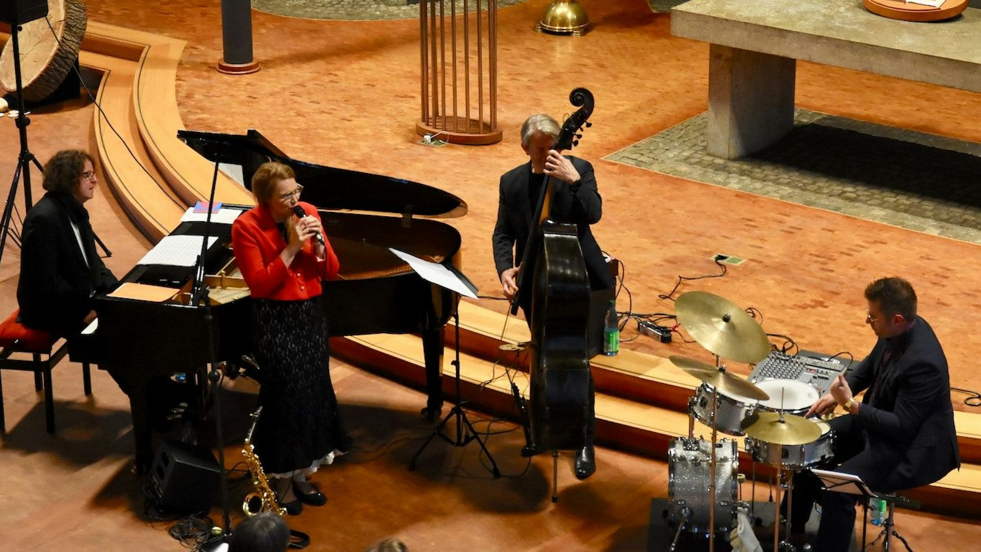 Männer und Frauen musizieren in einer Kirche. Sie spielen Bass, Schlagzeug und Klavier; eine Frau in einer roten Jacke singt.