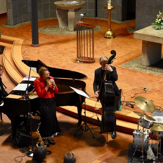 Männer und Frauen musizieren in einer Kirche. Sie spielen Bass, Schlagzeug und Klavier; eine Frau in einer roten Jacke singt.