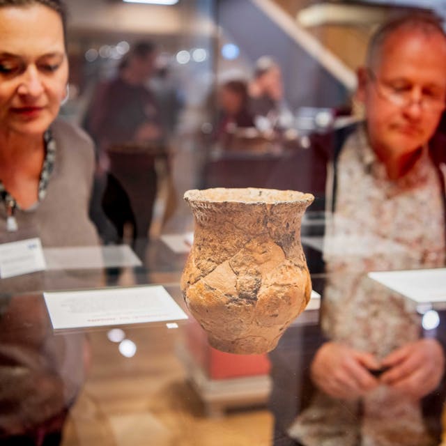 Das Bild zeigt einen Krug in einer Vitrine. Das Gefäß ist bei Großbüllesheim gefunden worden. Zwei Besucher schauen sich den Fund an.